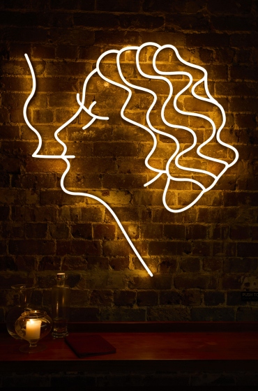 Neon outline of a woman's face in profile with wavy hair on a brick wall. Below, a candle flickers on a wooden surface, creating a cozy atmosphere.