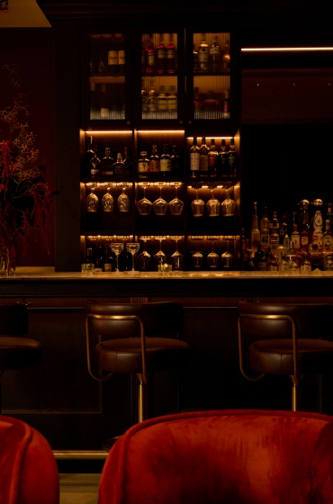 Dimly lit bar with a luxurious ambiance, featuring a wide array of bottles on shelves, velvet chairs, and bar stools. Warm and inviting atmosphere.