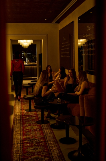 A dimly lit lounge with five women sitting on a couch, chatting and laughing. A person in a red jacket walks away on a patterned rug. Elegant chandeliers hang overhead, creating a warm ambiance.
