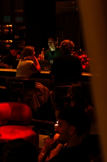 Dimly lit bar scene with people sitting at the counter, softly illuminated by red and orange hues. A plush red chair is in the foreground, creating a cozy atmosphere.