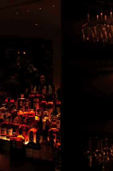 Dimly lit bar scene with a variety of illuminated liquor bottles on the counter, creating a warm glow. Glasses hang above, enhancing the cozy ambiance.