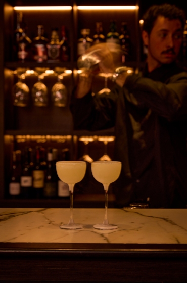Two elegant cocktails in coupe glasses on a marble bar, with a bartender in dim lighting shaking a mixer, surrounded by bottles on shelves.