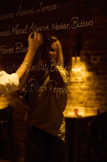 Person writing cocktail ingredients on a glass board in a dimly lit bar with warm lighting and brick walls.