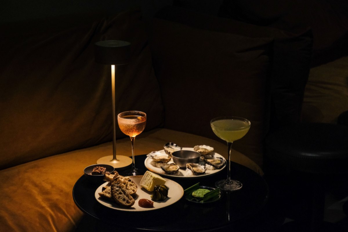 A dimly lit setting features a cozy table with oysters on a plate, a selection of appetizers, and two cocktails. The ambiance is elegant and intimate.