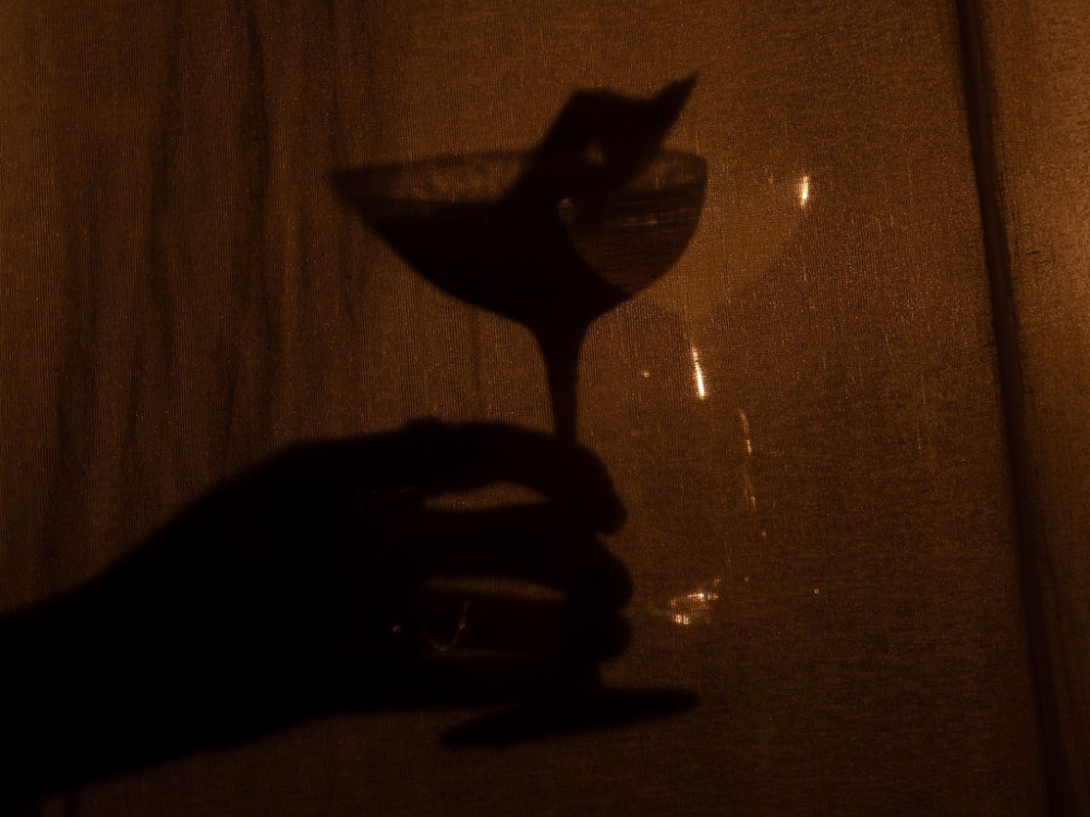 A silhouetted hand holds a cocktail glass against a sepia-toned background, creating a warm, mysterious ambiance with soft shadows.